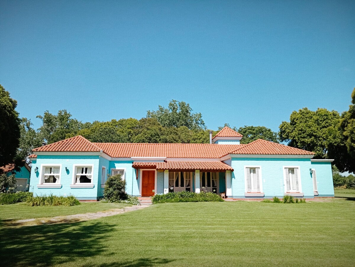 A single-story house features a light blue exterior with a terracotta-tiled roof. Large windows adorned with white frames provide ample natural light. Green grass surrounds the home, complemented by neatly trimmed bushes and a tree line in the background.