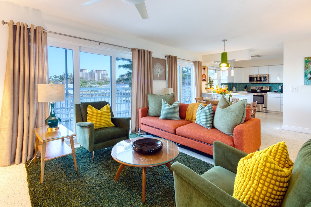 A vibrant living area features a modern sofa in orange, complemented by green accent chairs. A round coffee table sits on a plush green rug, while large windows showcase scenic waterfront views. Natural light fills the space, enhanced by tasteful decor and greenery.