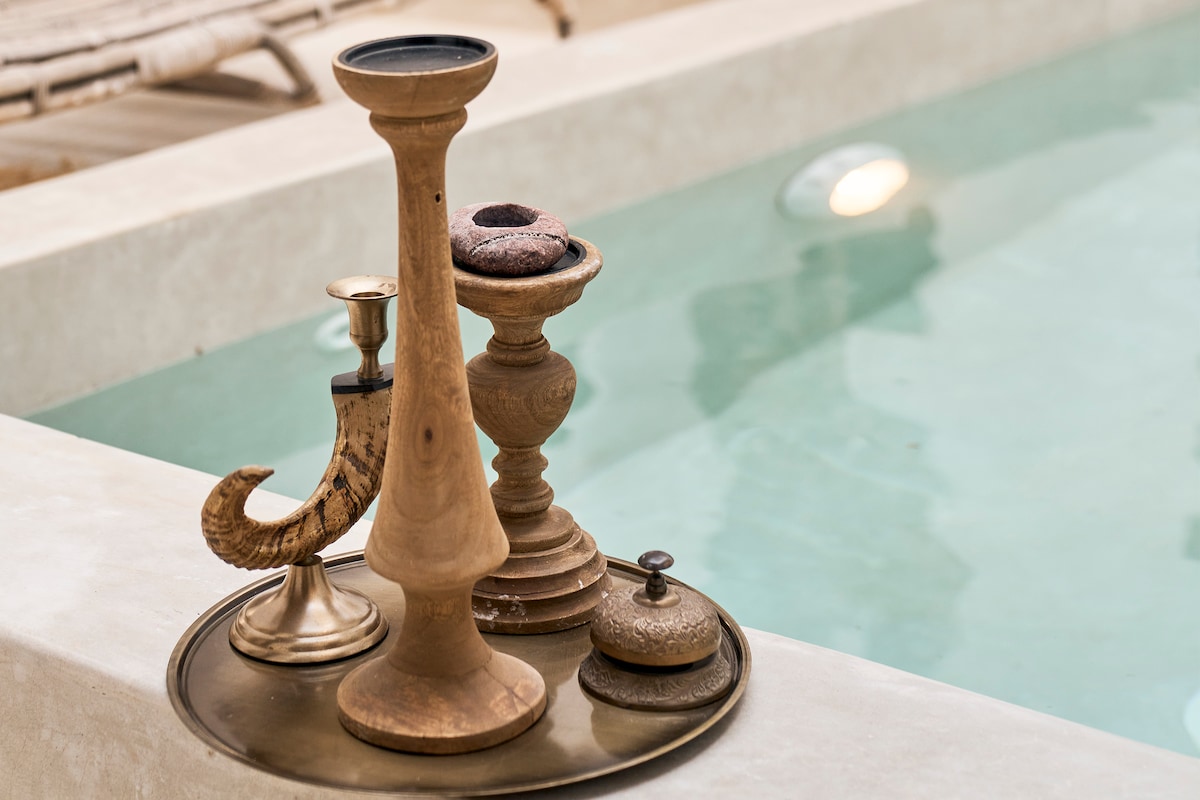 A decorative serving tray is positioned near a tranquil pool, featuring an assortment of candles in unique holders. The tray includes a wooden horn-shaped item, elegant candlesticks, and a small ornate container, all set against the calming blue water of the pool.
