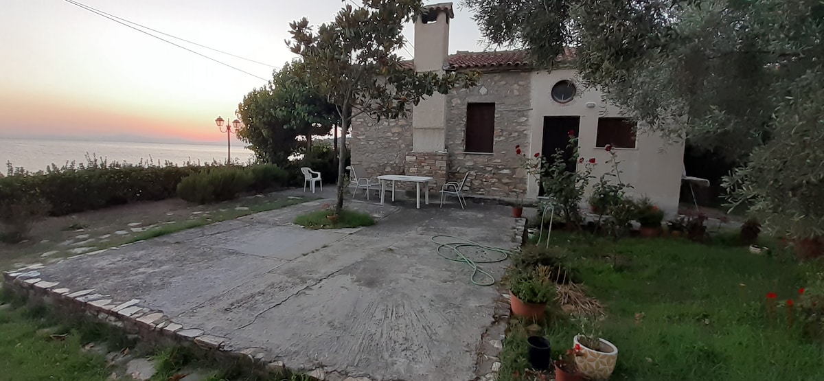 A stone cottage is set in a spacious outdoor area, featuring a concrete patio with garden plants and a table surrounded by chairs. Olive trees are visible, providing shade. The sunset casts a warm light across the scene, creating a calm ambiance.