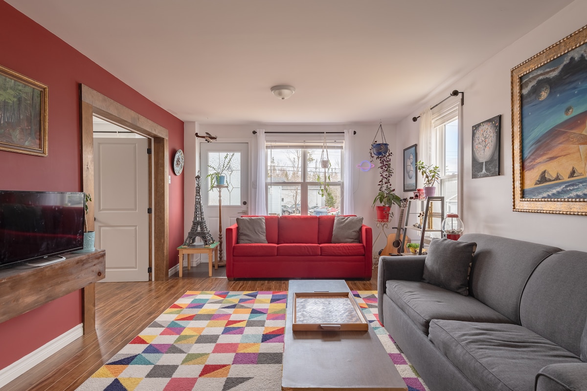 A cozy living room is presented, featuring two sofas—one red and one grey. A colorful, patterned rug lies on the floor, and art decorates the walls. Sunlight streams through large windows, with plants positioned in the corners, adding a touch of greenery to the space.