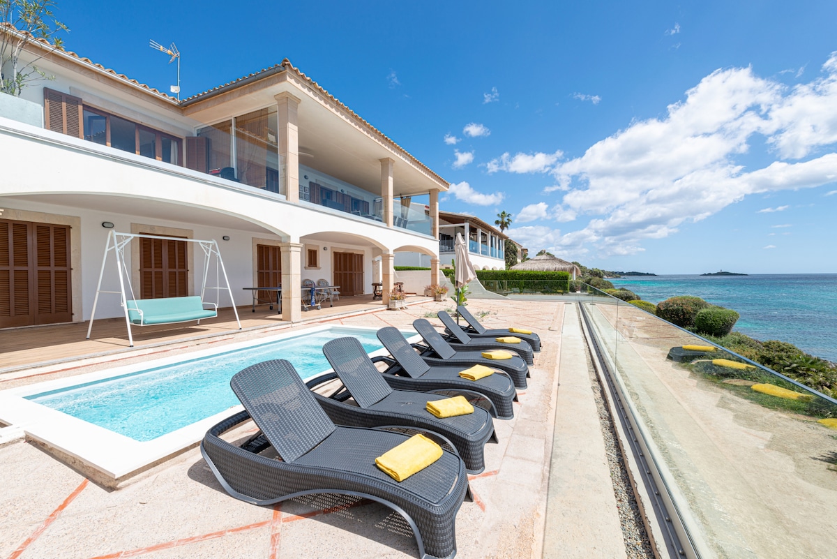 An outdoor terrace showcases a private pool bordered by six sleek sun loungers, each adorned with a bright yellow towel. A swing seat adds charm to the space, while a serene view of the bay and crystal-clear waters enhances the relaxing atmosphere.