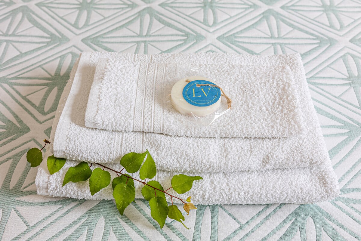 A neatly arranged stack of fluffy white towels is displayed on a patterned surface. A small, round bar of soap is placed atop the towels, accompanied by a sprig of green leaves, adding a touch of natural elegance to the setting.