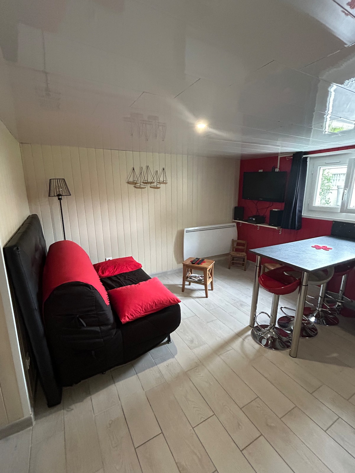 A compact living space is presented with a black couch featuring red accents and a small wooden chair. A dining table is positioned nearby, surrounded by sleek bar stools. A flat-screen TV is mounted on the wall, and natural light enters through a window adorned with dark curtains.