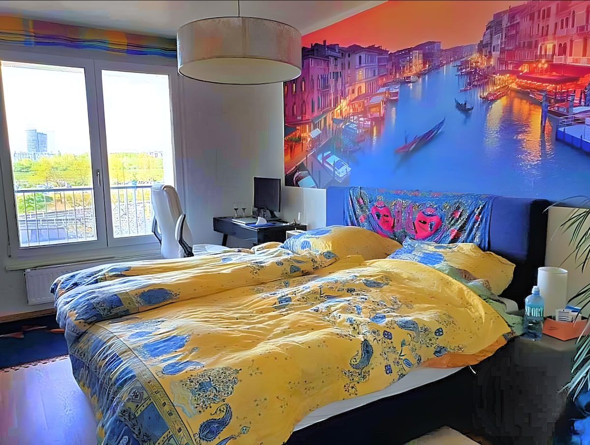 A spacious bedroom is presented with a striking mural of a vibrant canal scene. The large bed is dressed in a bright yellow and blue patterned duvet. A work desk is situated nearby, and natural light floods the room through two large windows.
