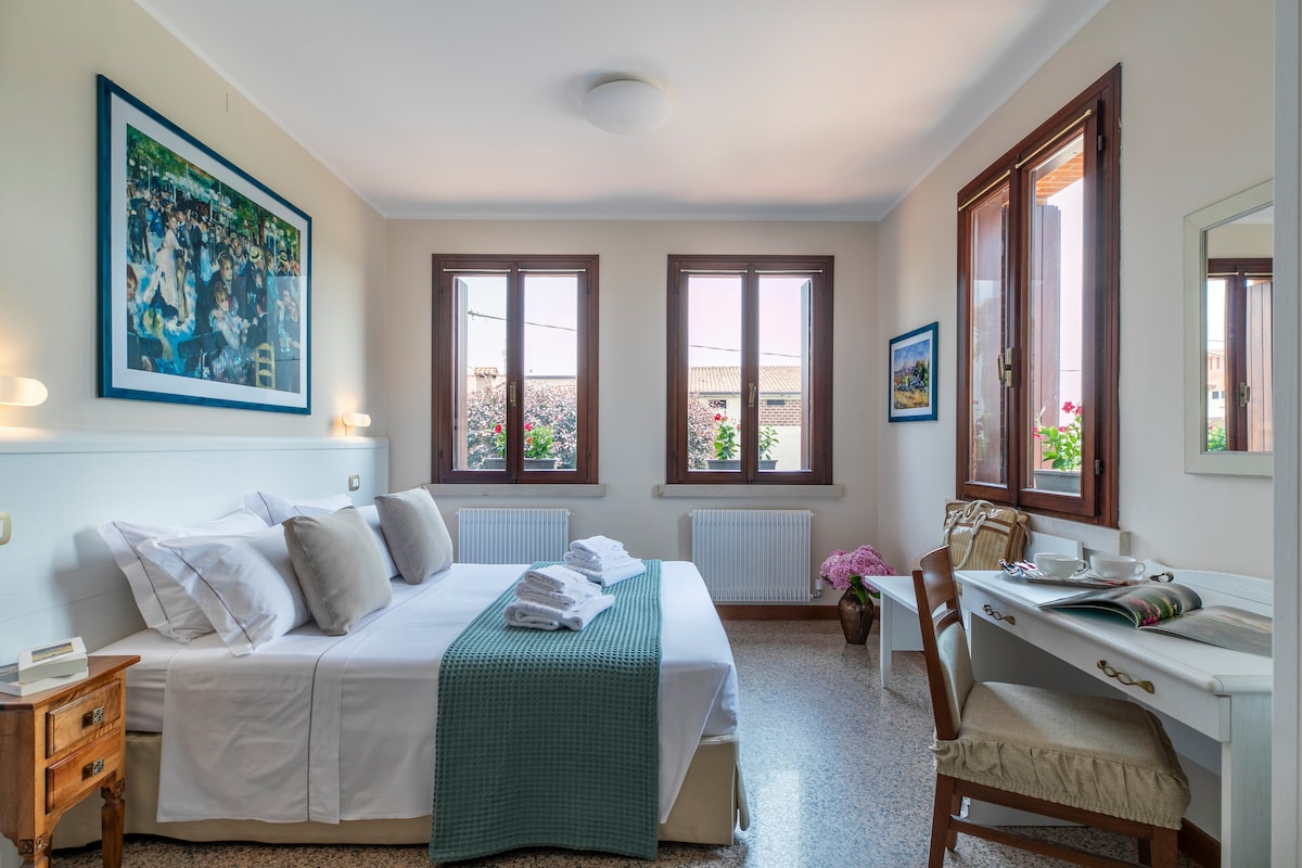 A spacious sleeping area features a comfortable bed adorned with neatly arranged white linens and decorative pillows. Natural light filters through two large windows with wooden frames, showcasing vibrant flower boxes. A small desk and mirror provide an inviting workspace.