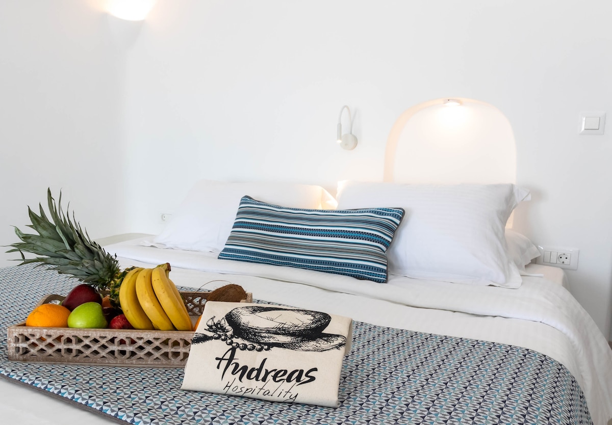 A comfortable bedroom is presented with a spacious bed dressed in crisp white linens and a textured throw. A decorative tray of fresh fruits is arranged on the bed, accompanied by a welcoming hospitality note. Soft lighting enhances the serene atmosphere.
