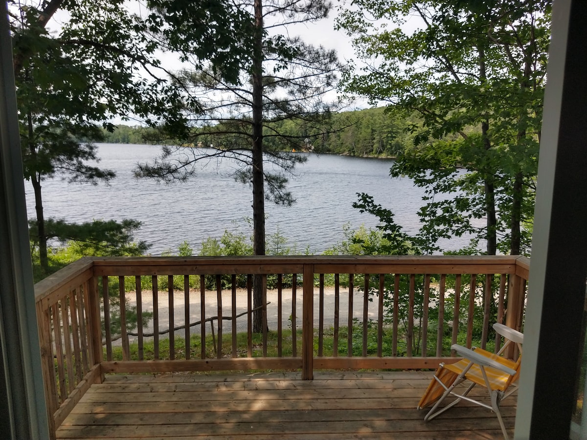 A wooden deck is shown with a simple chair, offering a view of a serene lake surrounded by trees. The calm water reflects the greenery, creating a peaceful setting. A pathway is visible in the background, leading alongside the lake.
