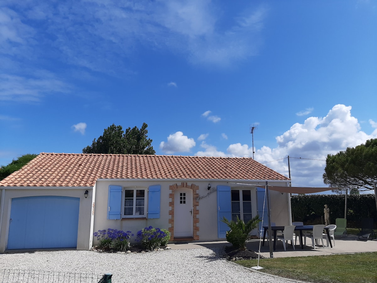 The exterior of a single-story house is visible, featuring blue wooden shutters and a light-colored façade. A gravel area surrounds the home, with outdoor furniture and potted plants nearby. A garage is attached to one side, and a clear blue sky is seen above.