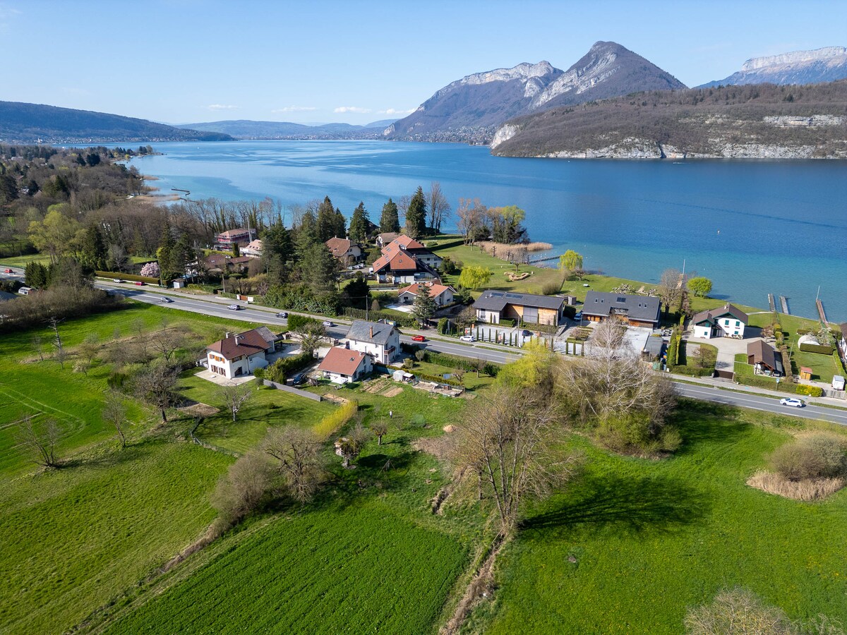An aerial view captures a serene landscape showing a diverse collection of homes near a tranquil body of water. Lush green fields and trees surround the area, with distant mountains visible under a clear blue sky.