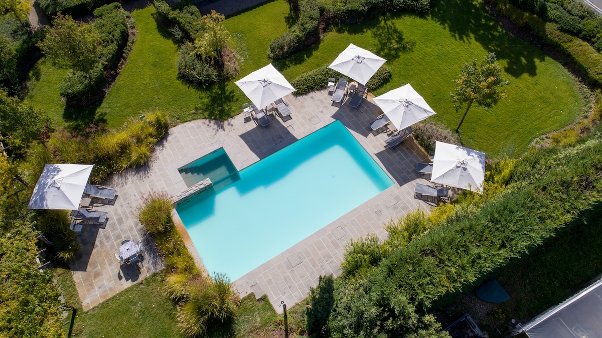 An aerial view displays a serene swimming pool surrounded by a well-manicured garden. Several lounge chairs are arranged under white umbrellas, creating shaded relaxation spots. Lush greenery borders the pool area, enhancing the tranquil setting.