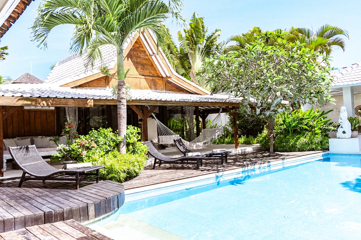 The outdoor space features a swimming pool surrounded by tropical greenery, with sun loungers positioned nearby. A covered lounge area with hammocks is included for relaxation. A stone sculpture and lush plants enhance the serene ambiance, inviting guests to enjoy the tranquil environment.