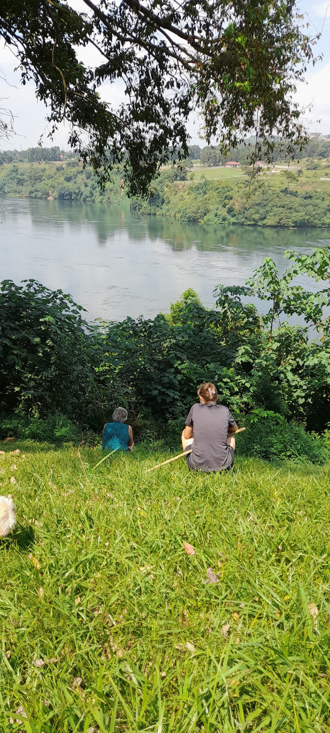 Two people are seated on a grassy slope overlooking the River Nile. Lush greenery surrounds them, and the water reflects the blue sky. The peaceful landscape invites relaxation, with trees framing the serene view of the river and nearby hills.