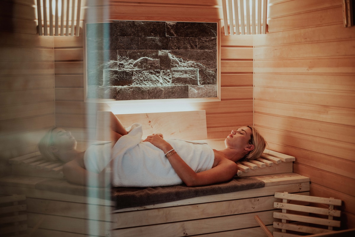 A wooden sauna interior features slatted benches and stone accents. Soft lighting creates a soothing ambiance. A person reclines peacefully, draped in a white towel. The walls are lined with natural wood, enhancing the tranquil atmosphere.