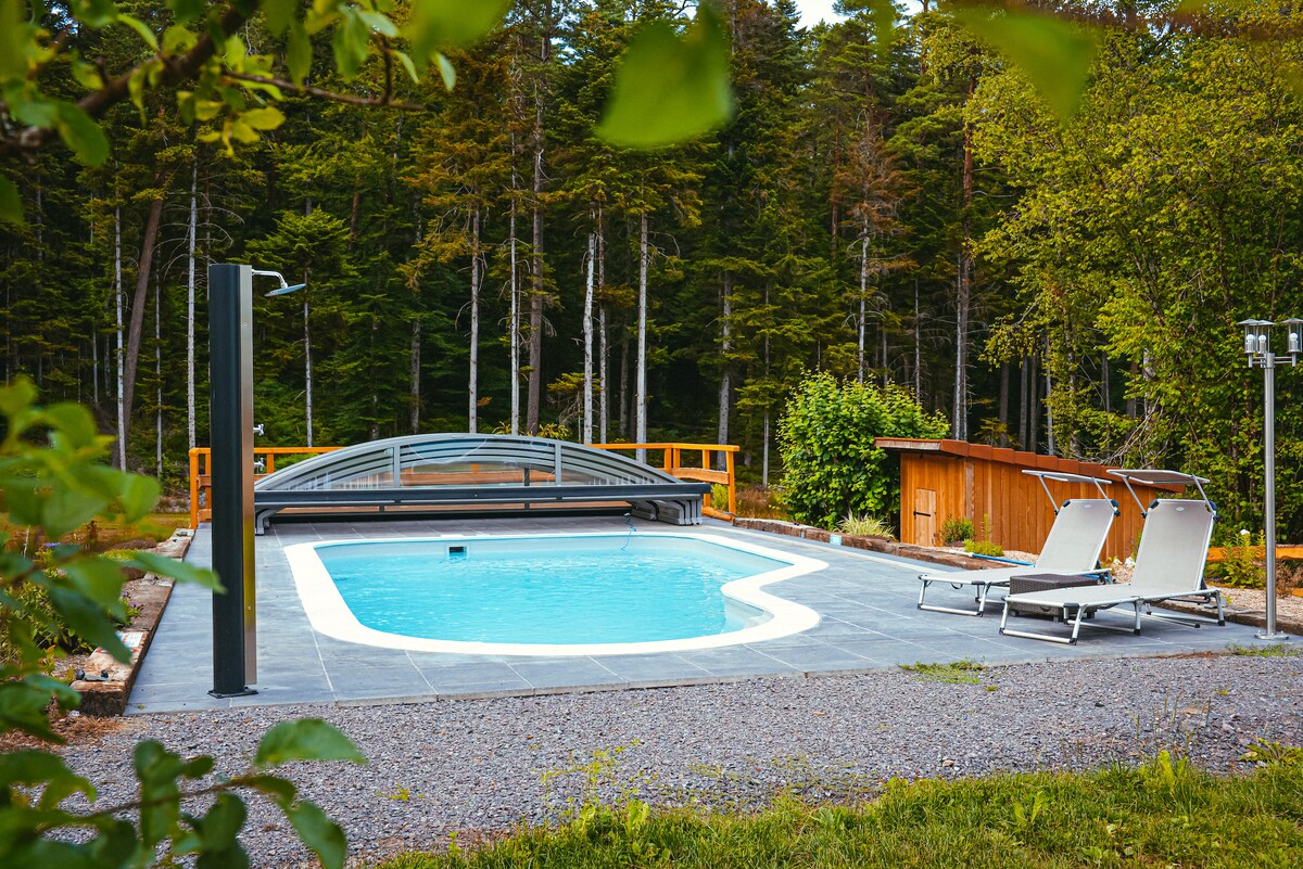 A serene outdoor space features a private pool surrounded by a gravel patio. Two lounge chairs are positioned poolside, and a wooden shed is visible nearby, all set against a backdrop of lush forest greenery.