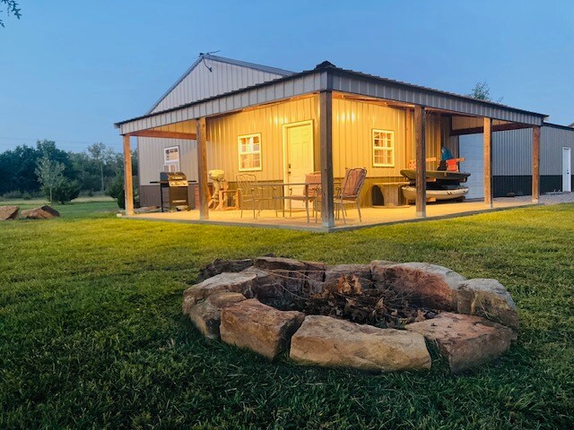 A covered porch extends from a metal structure, warmly lit in the evening. A fire pit made of stone is positioned in the foreground, surrounded by grassy areas. Additional seating, a grill, and a boat can be seen on the porch, enhancing the outdoor living space.