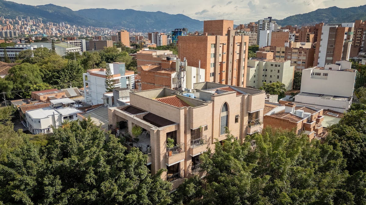 An aerial view showcases a large building surrounded by greenery, with a mountainous backdrop. The structure features a spacious layout with various rooflines and an arched window, indicating a blend of modern and traditional architecture within an urban setting.