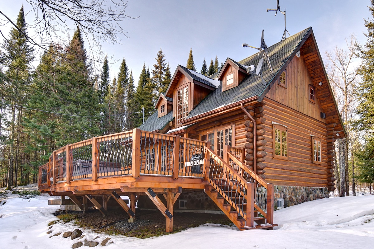 A charming wooden house is surrounded by trees, featuring a spacious deck with wooden railings. The exterior showcases a blend of logs and stone, with multiple windows that invite natural light. Snow is visible on the ground, highlighting the serene natural setting.