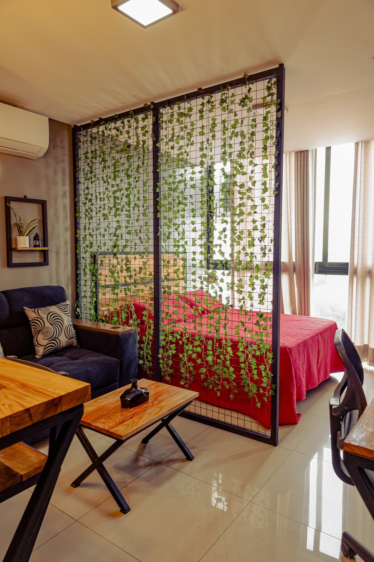 A modern studio is partitioned by a decorative green wall, offering a sense of privacy. A spacious bed dressed in red linens is visible, alongside a wooden table and two seating areas. Natural light flows in through large windows, enhancing the open space.