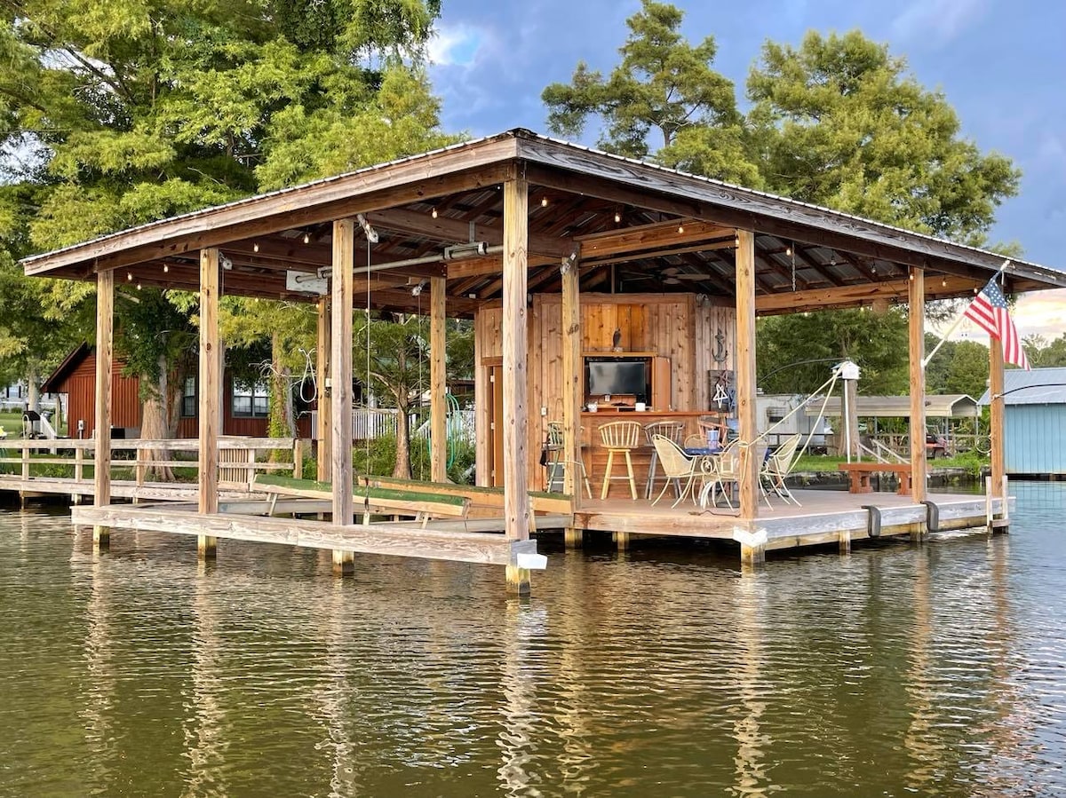 A spacious outdoor deck with wooden beams and flooring is illustrated, featuring a dining area and a lounge space. A television is mounted for viewing enjoyment. An American flag is visible on the edge, surrounded by water and trees, enhancing the waterfront setting.
