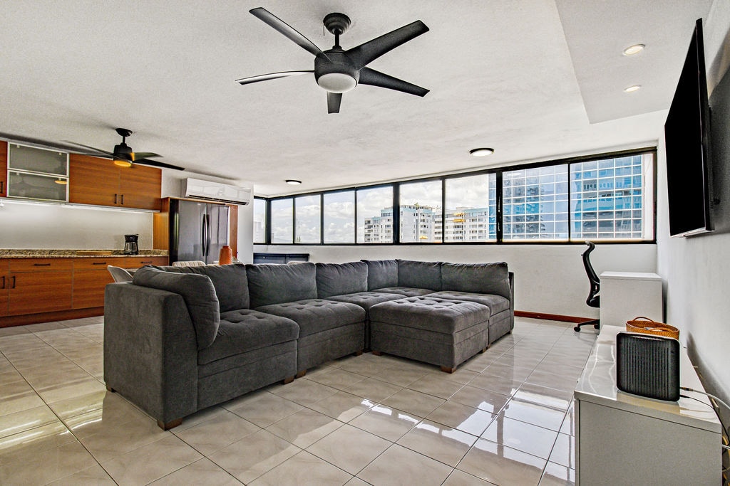 A spacious living area is highlighted by a large sectional sofa and a flat-screen TV mounted on the wall. Natural light flows through large windows, offering views of the surrounding buildings. Glossy tile flooring reflects the light, enhancing the airy feel of the space.