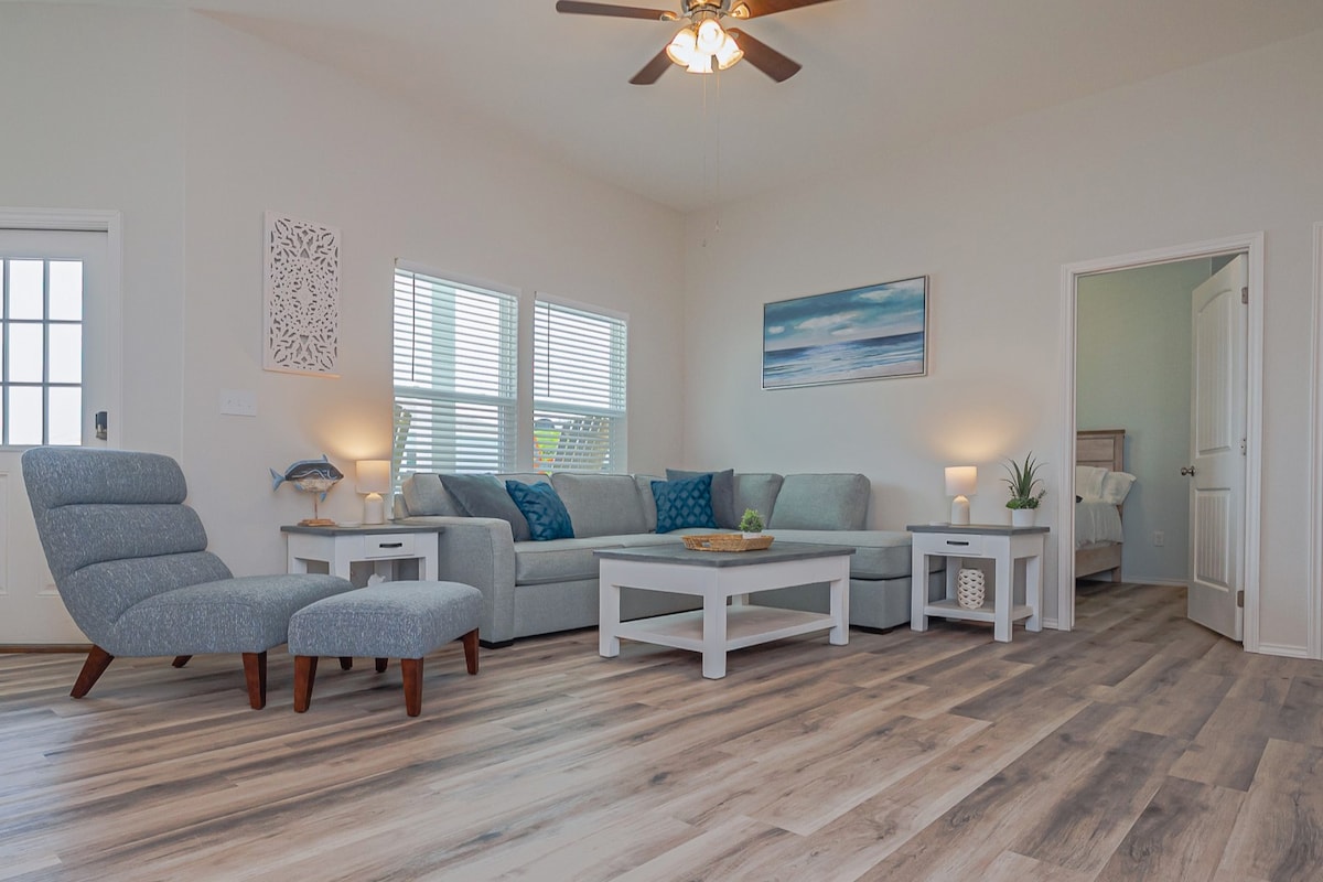 A spacious living area is highlighted by light-colored walls and wood laminate flooring. A comfortable grey sofa with decorative pillows faces a coffee table, while a lounge chair is positioned nearby. Natural light filters through multiple windows, creating a bright and inviting space.