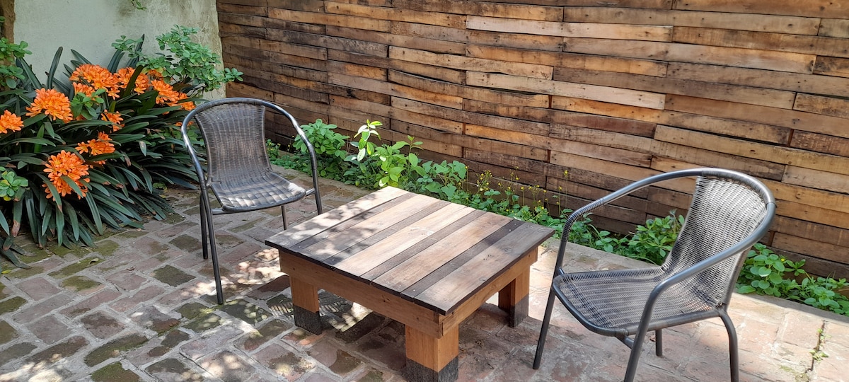 A cozy outdoor seating area features two black chairs arranged around a wooden coffee table. A backdrop of natural wood paneling is complemented by vibrant orange flowers and lush greenery, creating a tranquil space for relaxation.