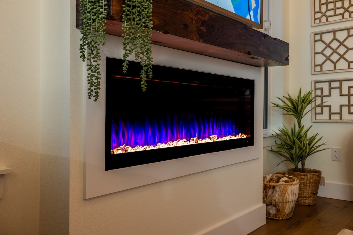 An electric fireplace is prominently displayed on a modern mantel, featuring vibrant blue and purple flames that create a soothing ambient glow. Live plants are positioned beside the fireplace, adding a touch of greenery to the warm decor.