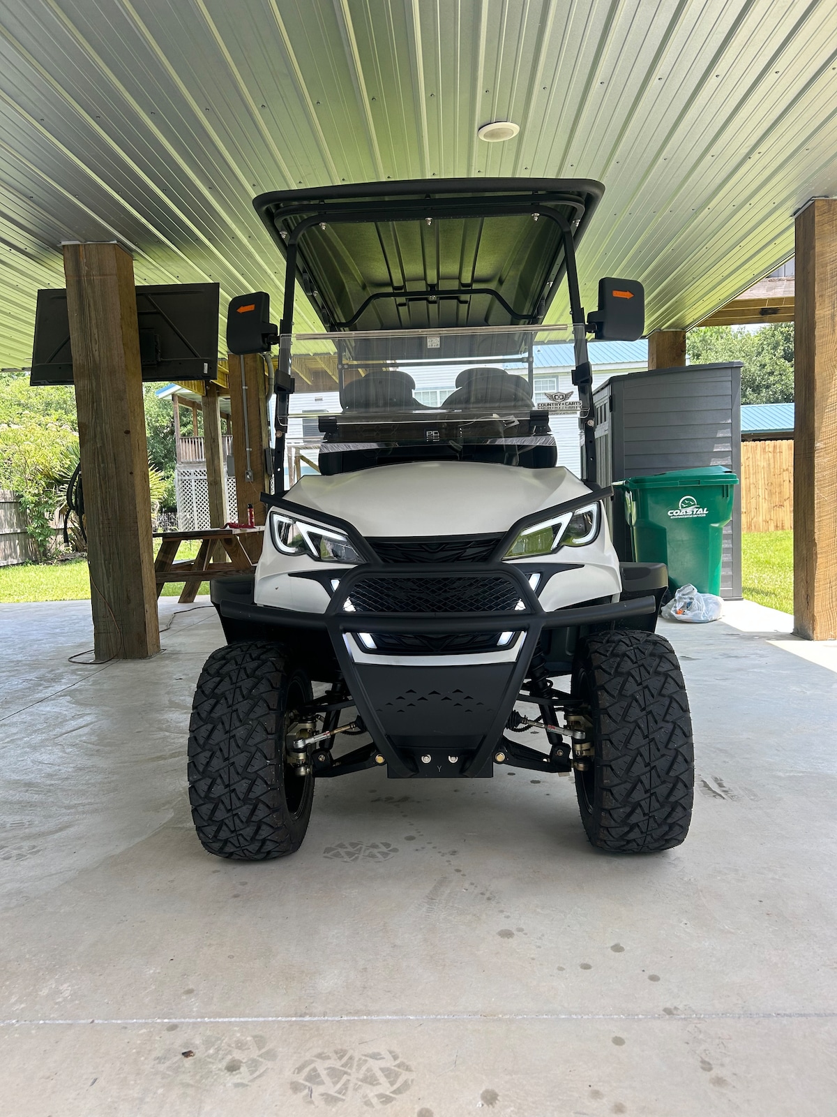 A golf cart is positioned on a concrete surface, featuring a sleek design with large wheels and a protective windshield. The cart is partially shaded by an overhead canopy, with an outdoor television visible in the background and greenery peeking through a nearby structure.