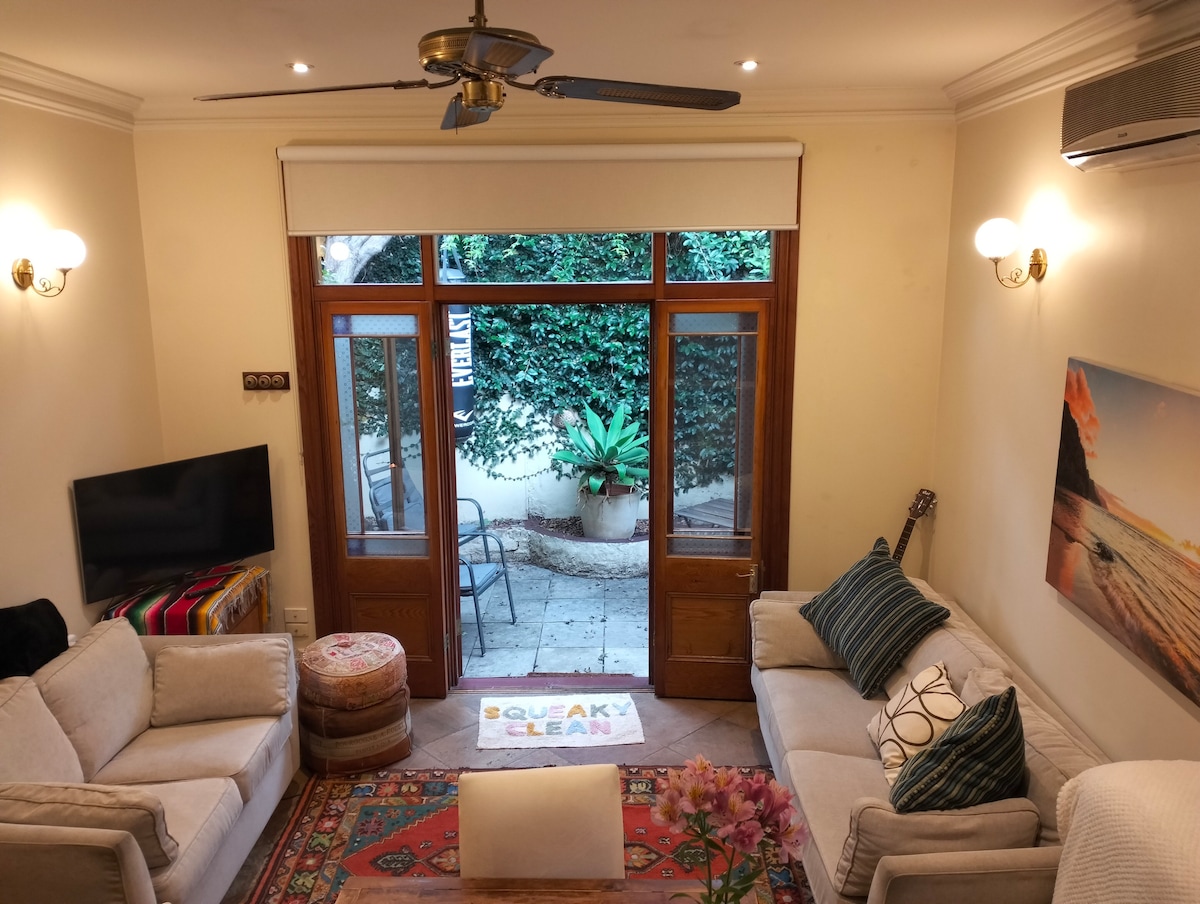 A spacious living area features light-colored sofas and a colorful area rug, complemented by a coffee table. Large glass doors open to a private patio adorned with plants. The space is illuminated by wall sconces, creating a bright and welcoming environment.