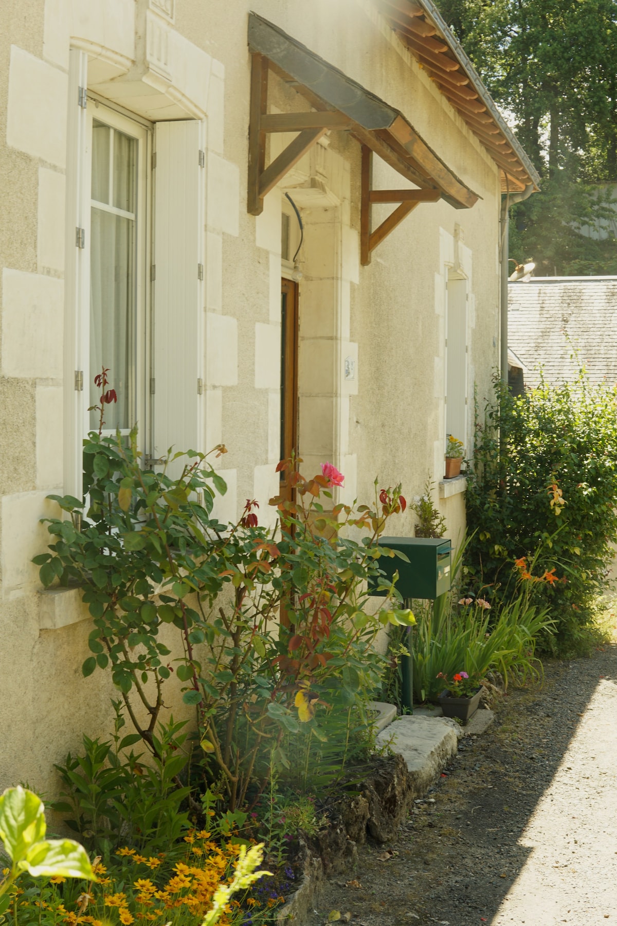 The exterior of a renovated house is shown, featuring a welcoming entrance with a small awning. Vibrant flowers and greenery decorate the pathway, creating an inviting appearance. The light stone facade contrasts with the surrounding flora, enhancing the charm of the property.