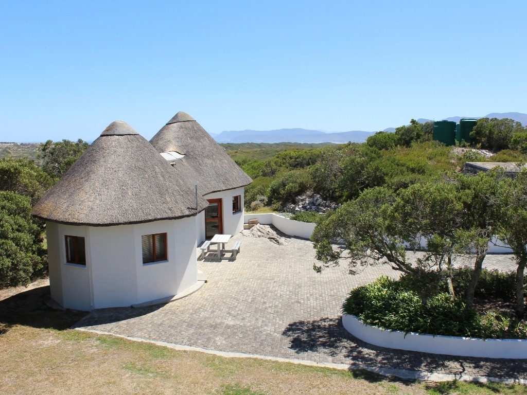 A semi-detached bungalow features a thatched roof and white exterior. A stone patio with a picnic table is visible in front, surrounded by natural greenery. Rolling hills and a clear blue sky create a serene backdrop, enhancing the sense of remoteness.