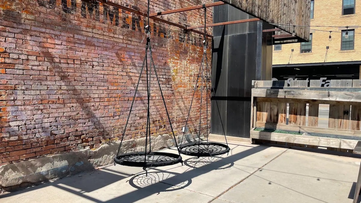 A cozy outdoor area features two hanging swing seats adorned in black metal, positioned against a rustic brick wall with a warm hue. A concrete pathway runs beneath, with a wooden structure visible in the background. String lights above add a subtle ambiance.