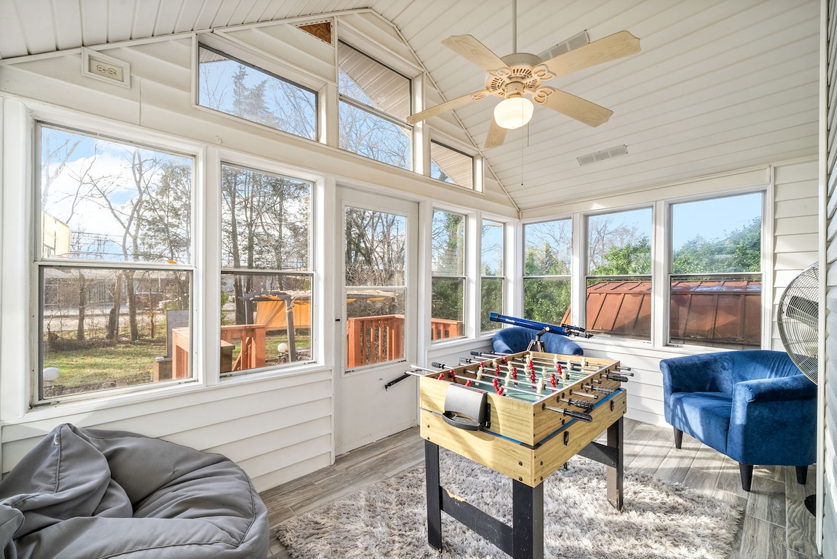 A sunlit sunroom features a foosball table at its center, surrounded by large windows that invite natural light. A blue armchair and a cozy bean bag offer comfortable seating options, with a ceiling fan positioned above for added airflow.