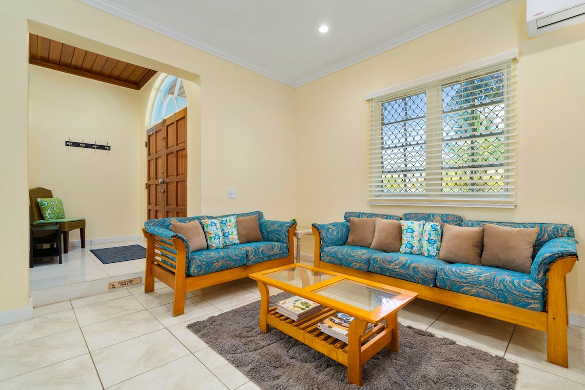 A welcoming living area features two wooden sofas adorned with colorful cushions, complemented by a glass coffee table. Natural light enters through the windows with privacy screens, illuminating the tiled floor. A small entryway with a bench is visible in the background.