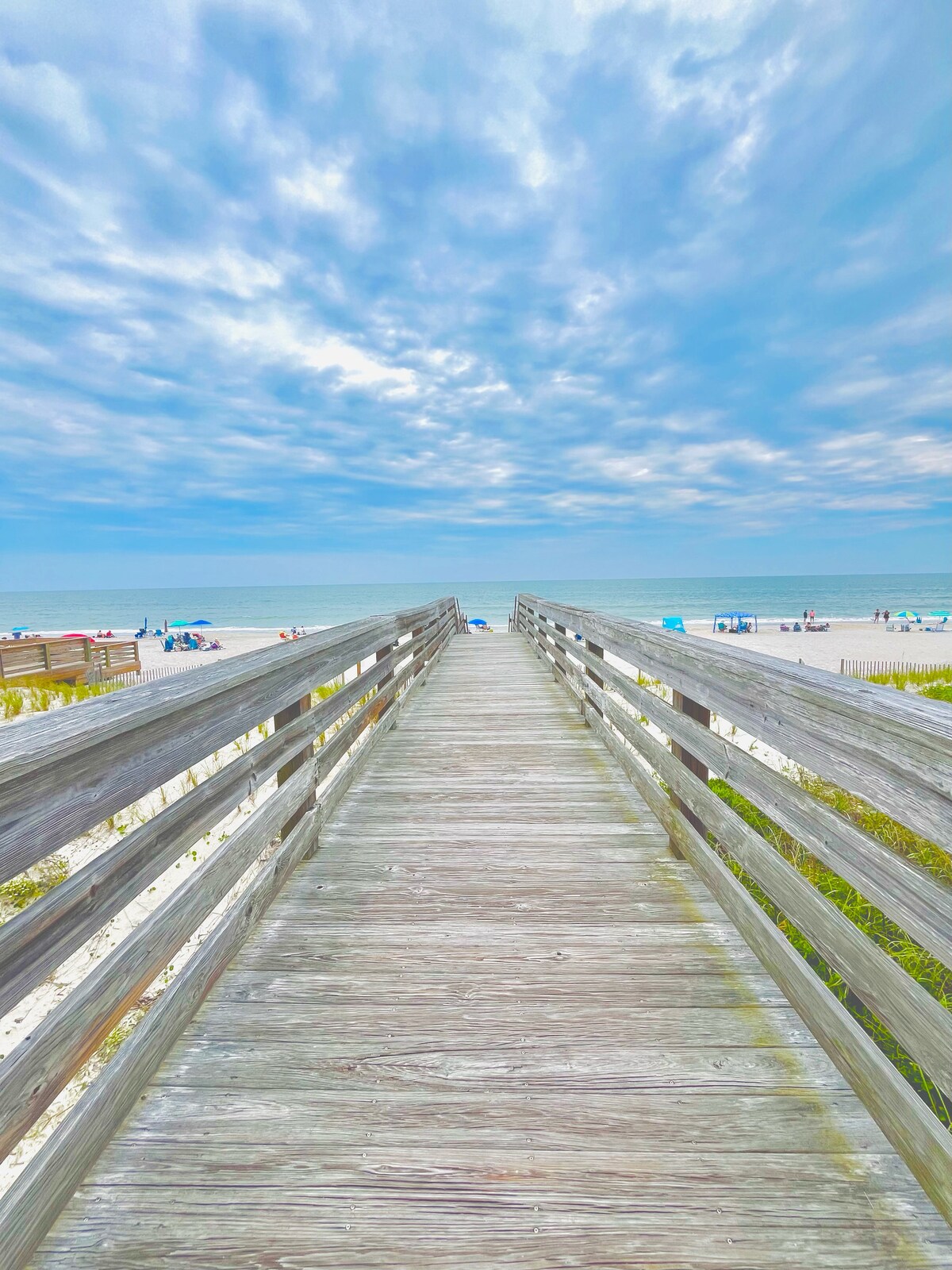 A wooden boardwalk leads towards the beach, flanked by grassy areas. Sunbathers and umbrellas dot the sandy shore, with calm waves lapping at the shoreline. A cloudy sky stretches overhead, complementing the serene atmosphere of the coastal scene.