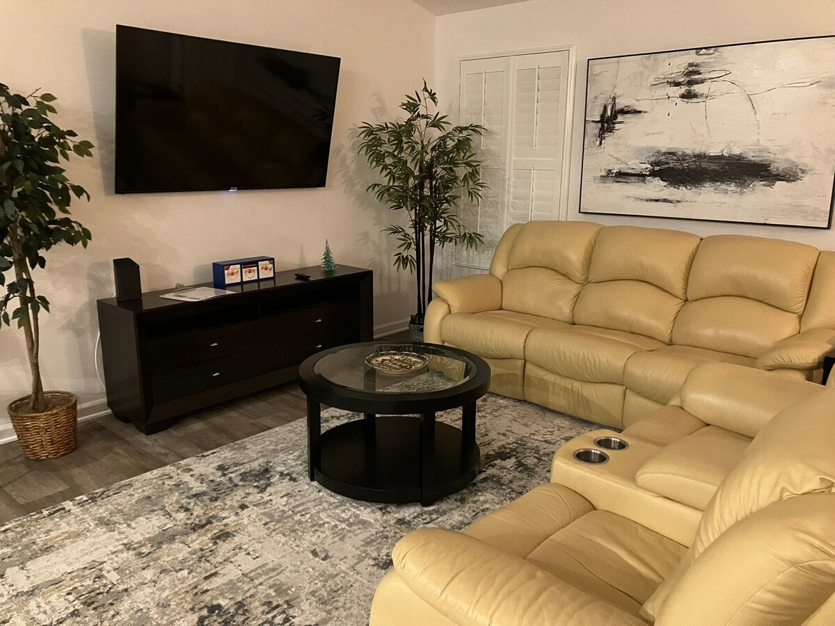 A comfortable living area features a large flat-screen television mounted on the wall. The seating includes stylish golden leather couches, arranged around a round coffee table on a softly patterned area rug. A potted plant in the corner adds a touch of greenery.