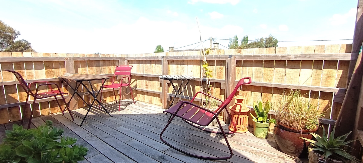An elevated terrace area is visible, featuring wooden decking and surrounded by wooden fencing for privacy. Two pink chairs and a small table are arranged, along with potted plants adding greenery. Natural light brightens the space, creating a welcoming outdoor environment.