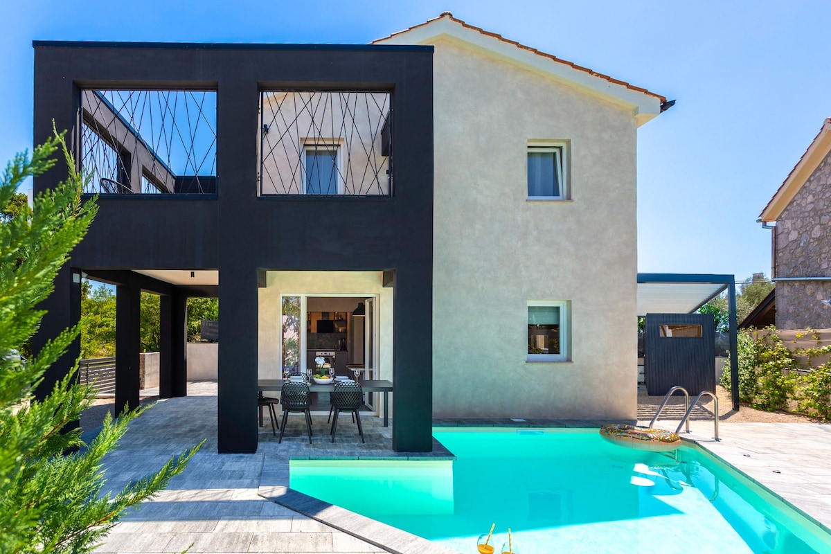 The villa presents a two-story structure with a modern facade, featuring large windows. A spacious outdoor area includes a pool, accompanied by a dining set under the shade of a covered porch. Lush greenery surrounds the property, enhancing the serene environment.
