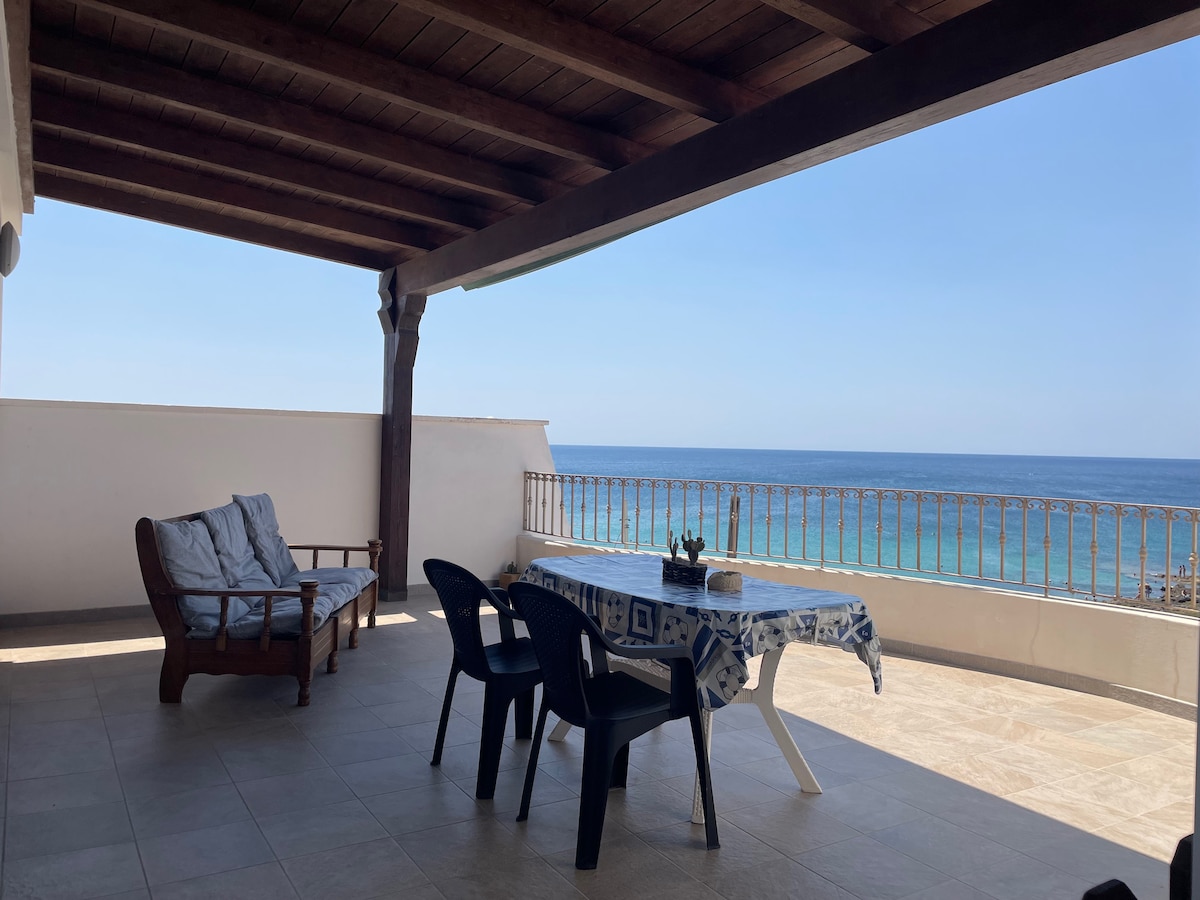 A spacious terrace provides an unobstructed view of the sea. Comfortable seating is arranged alongside a dining table, covered with a tablecloth. The flooring is tiled, and wooden beams support the overhead structure, contributing to a relaxed outdoor atmosphere.