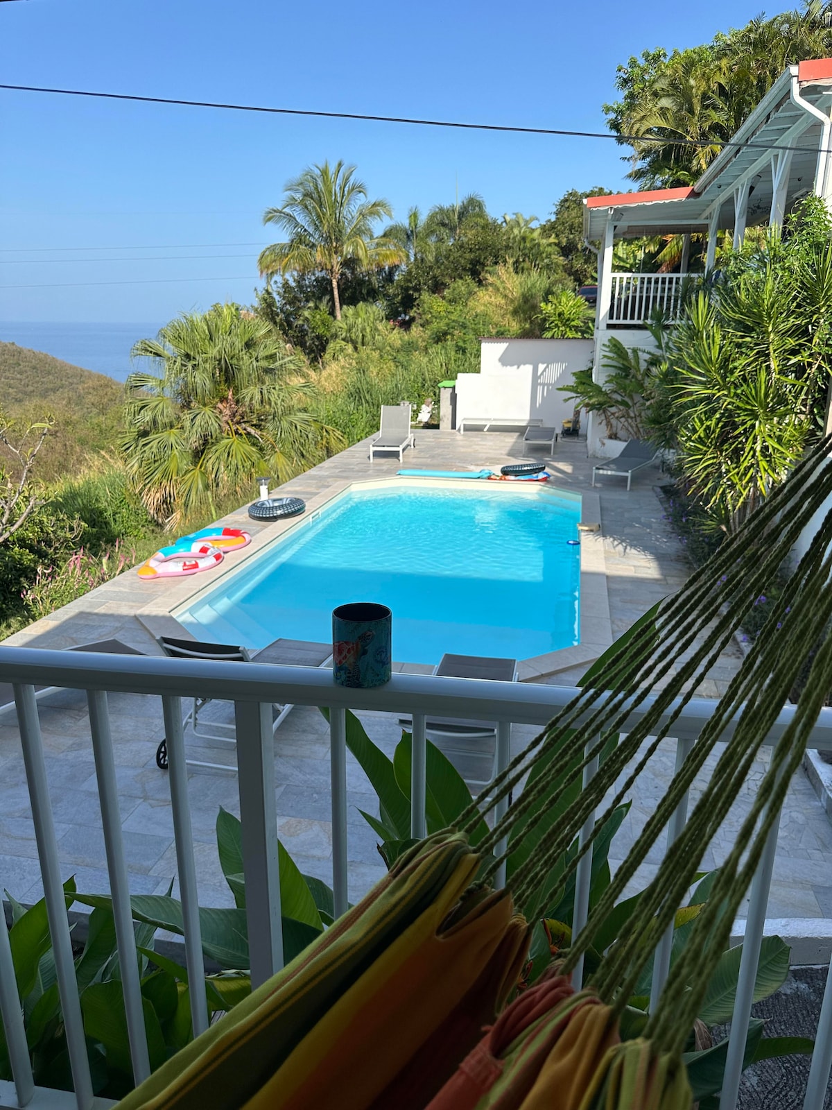 A tranquil outdoor space features a clear blue pool surrounded by lush greenery and sun loungers. Palm trees provide shade nearby, while colorful floats rest on the water's surface. A hammock hangs in the foreground, suggesting a relaxed atmosphere for guests.
