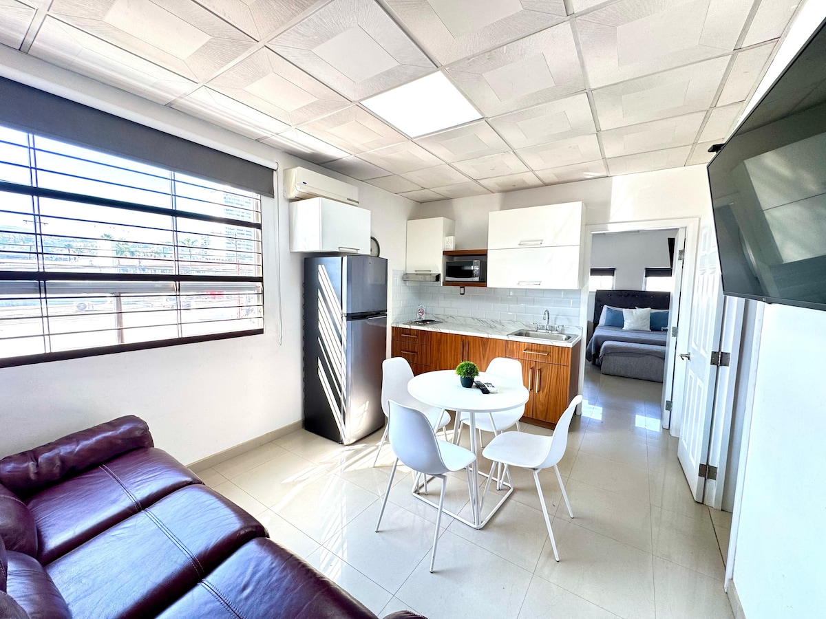 A bright open-plan living area features a modern kitchen with white cabinets and stainless steel appliances. A round dining table with four chairs sits adjacent to a cozy leather sofa. Natural light filters through the large window, highlighting the tiled flooring.