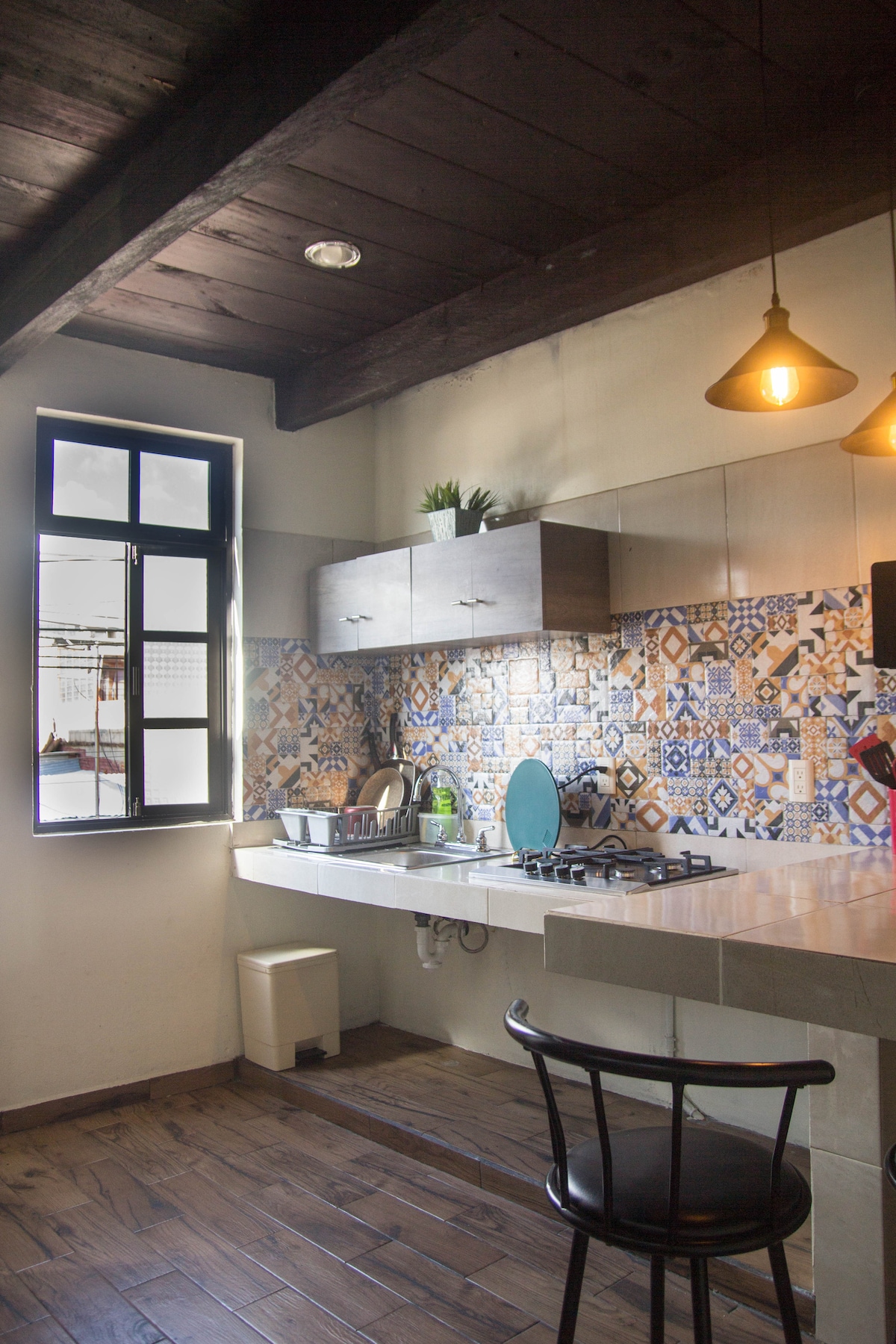 The kitchen features a modern design with colorful tiled backsplash and a large counter space. Natural light pours in through a window, illuminating a stovetop, sink, and various kitchen essentials. A black bar stool complements the minimalist decor, creating a functional cooking area.