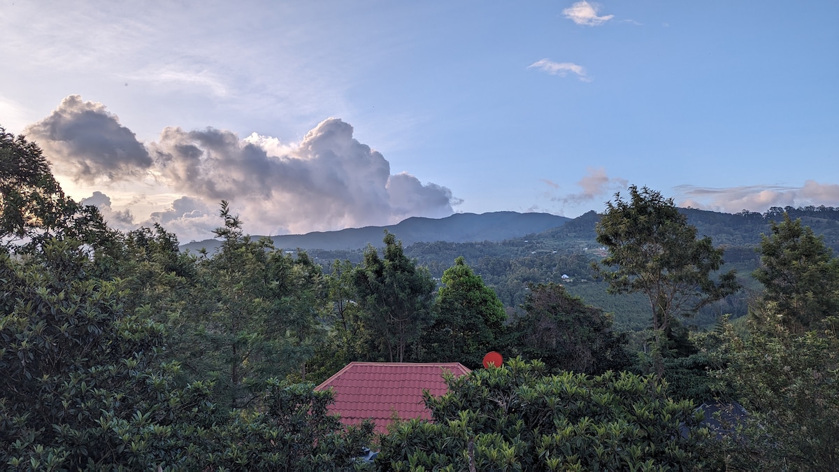 A serene view is presented, featuring distant mountains and a sky with soft clouds. Lush greenery surrounds a red-roofed house, while various trees create a natural frame, emphasizing the peaceful ambiance of the hillside setting.