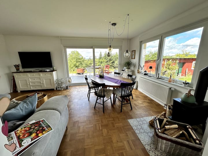 Wohnzimmer mit einer gemütlichen Couch, einem Esstisch und einem wunderbaren Panoramablick in die Natur. Freier Zugang zur Terrasse. Zudem steht im Wohnzimmer ein kleiner Gussofen der Marke Jotul, für den Brennholz bereitgestellt ist.