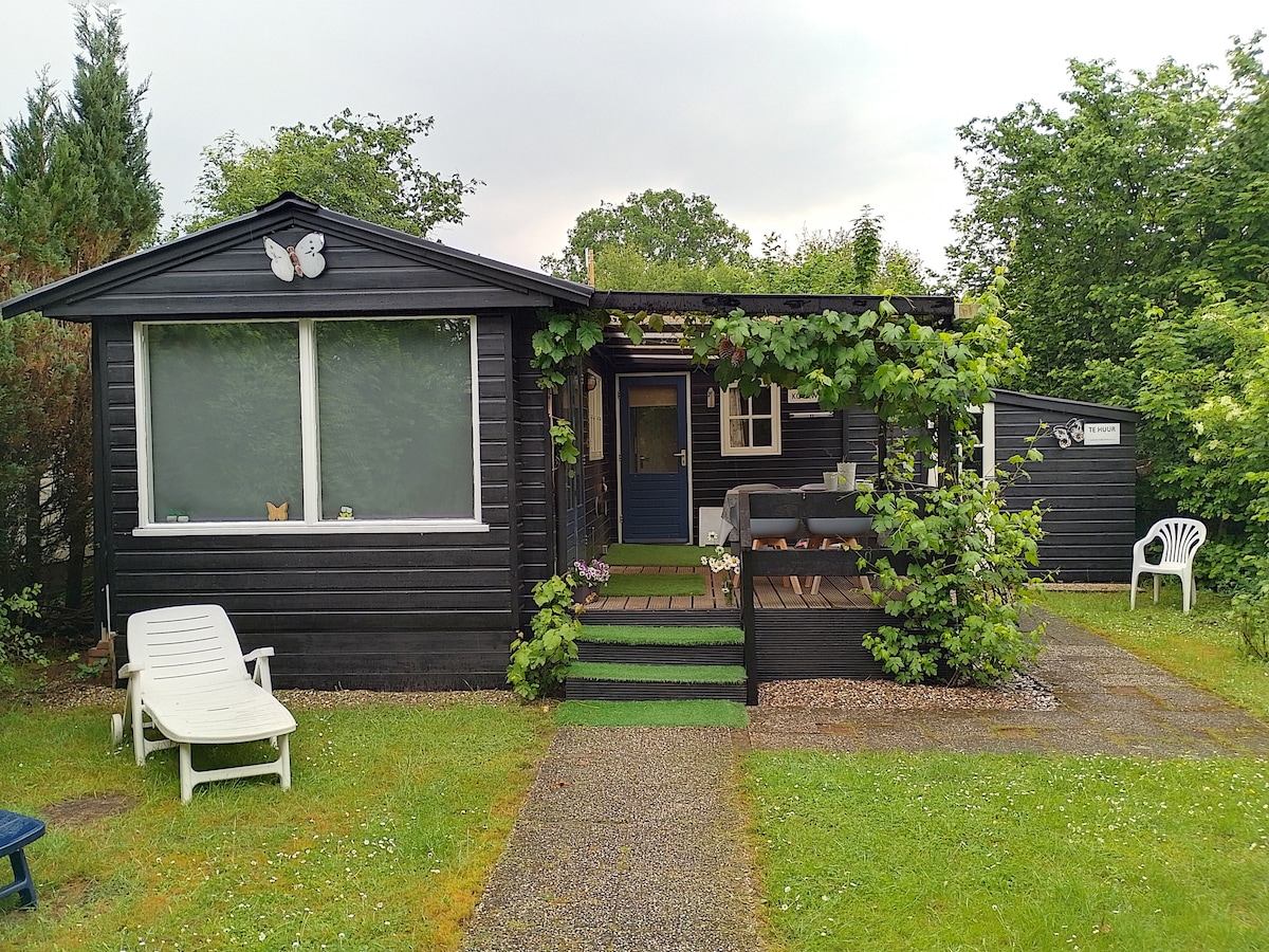 A small black cottage is surrounded by greenery, featuring a front deck with a seating area. A large window brings in natural light, and decorative elements such as butterflies add charm. The pathway is lined with gravel, leading to a well-maintained lawn.