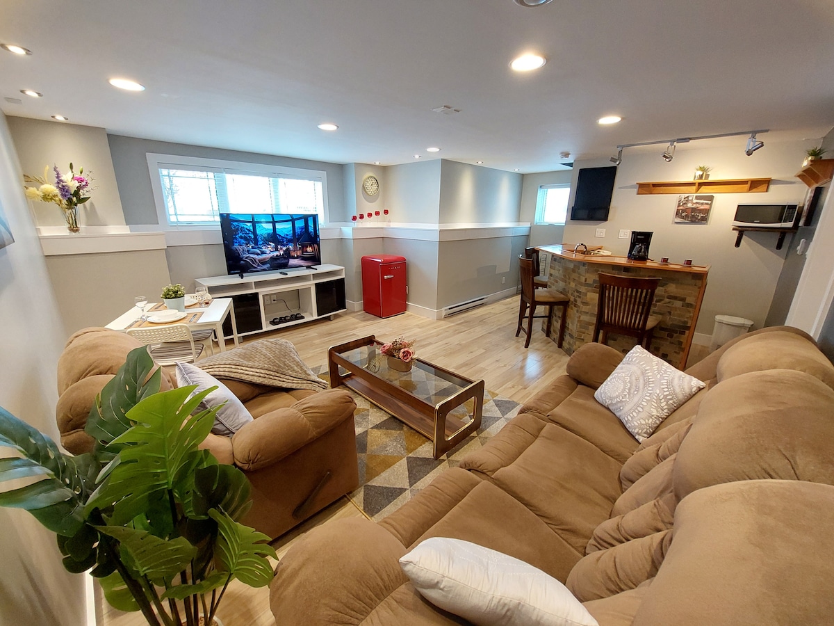 A comfortable living area features a spacious sectional sofa, a coffee table, and a Samsung television displaying a scenic image. Natural light enters through the windows, while a small bar area with stools is visible in the background, alongside a compact red refrigerator.