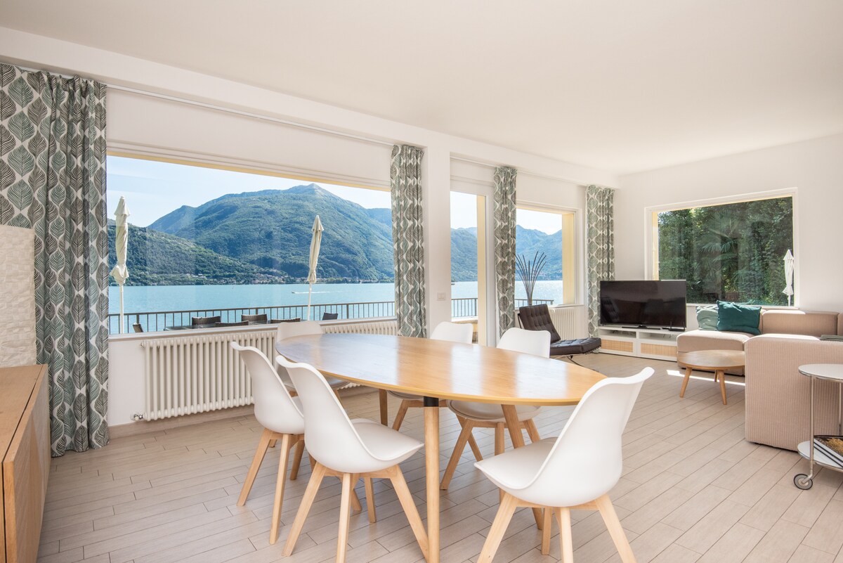A bright and airy living area features large windows with lake views, inviting natural light. A wooden dining table with white chairs is positioned in the center, surrounded by modern furnishings. The space showcases a neutral color palette and indoor plants, offering a serene atmosphere.