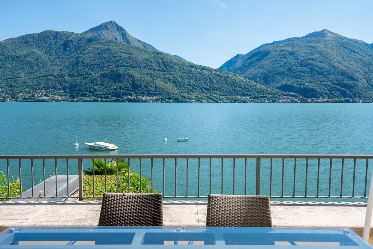 A view of the tranquil lake is captured, framed by distant mountains. The calm water reflects the clear blue sky, while two small boats are visible on the surface. A terrace with a glass table and woven chairs is positioned in the foreground.