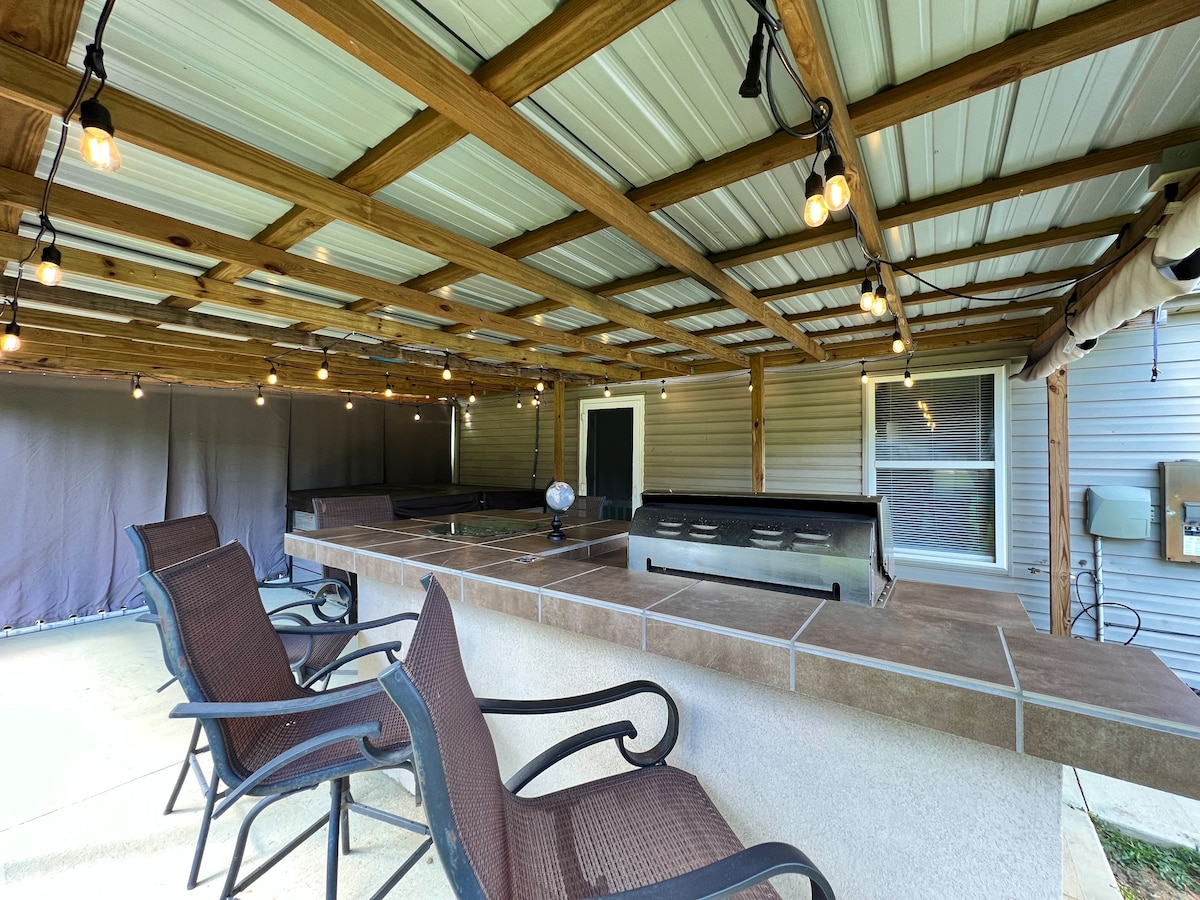 A covered patio area features a spacious dining island with a grill, surrounded by four chairs. Soft lighting hangs from the wooden beams, creating a cozy ambiance. The patio is designed for outdoor dining and relaxation, providing a seamless connection to the backyard.